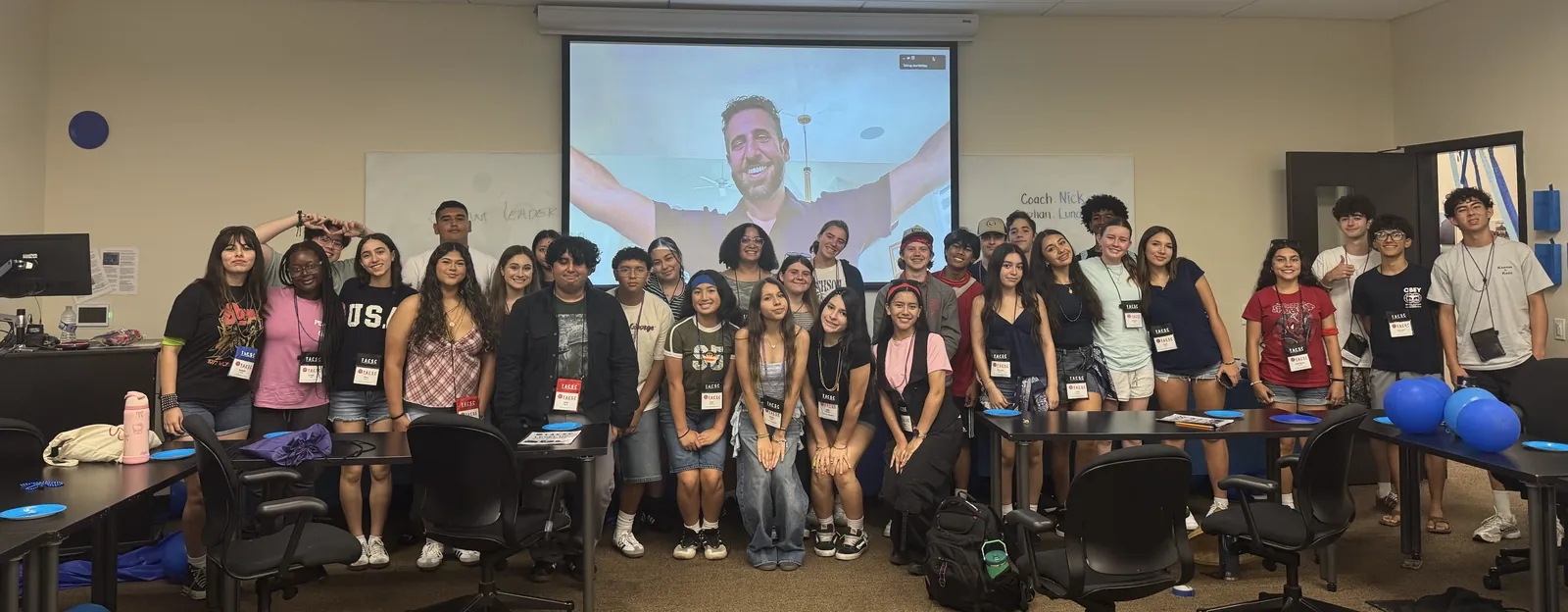 Thirty-five high school students standing together at a TACSC leadership workshop, with the facilitator joining on the screen behind them.